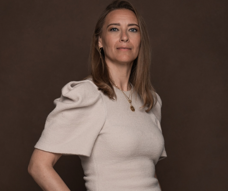 En kvinde med glat, lysebrunt hår iført en lys beige, struktureret kjole med pufærmer står på en mørkebrun baggrund og ser selvsikkert ind i kameraet. Hun bærer en guldhalskæde og øreringe.